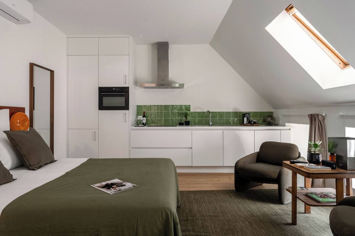Un dormitorio con cama, escritorio, silla y una kitchenette al fondo.