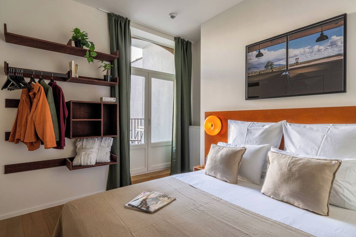 Habitación de hotel con cama, televisor de pared, ventana con cortinas y estante con ropa y plantas.
