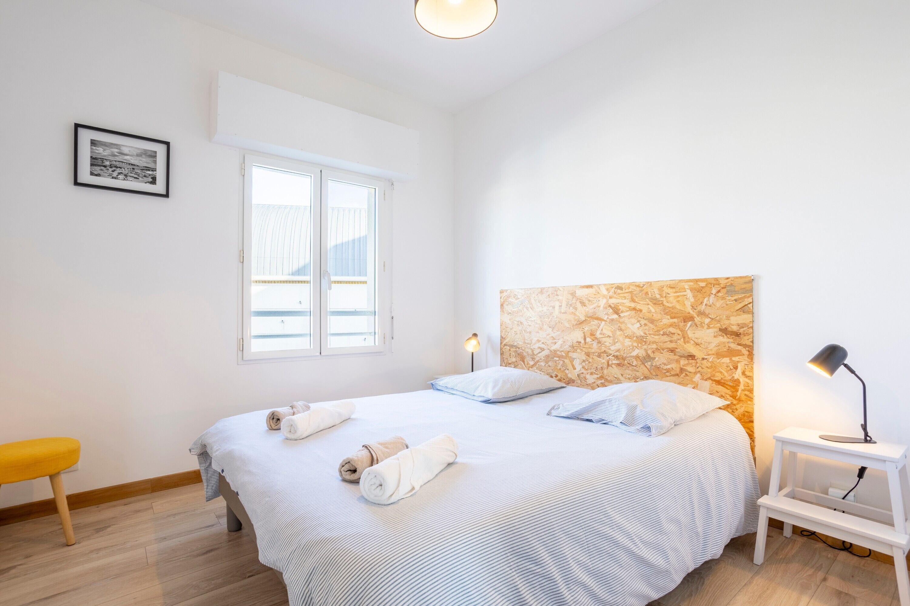 Une chambre à coucher avec un lit, une table de chevet avec une lampe, une fenêtre et un tableau encadré au mur.