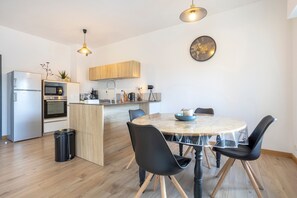 A modern kitchen with a dining area, wooden cabinets, a built-in oven, and a round table with chairs. - L'Urania – Stay in the Heart of Concarneau (Concarneau)