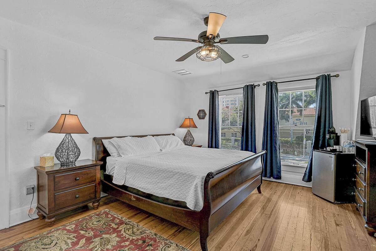 A bedroom with a wooden bed, a ceiling fan, a nightstand with a lamp, a dresser, and a window with curtains.
