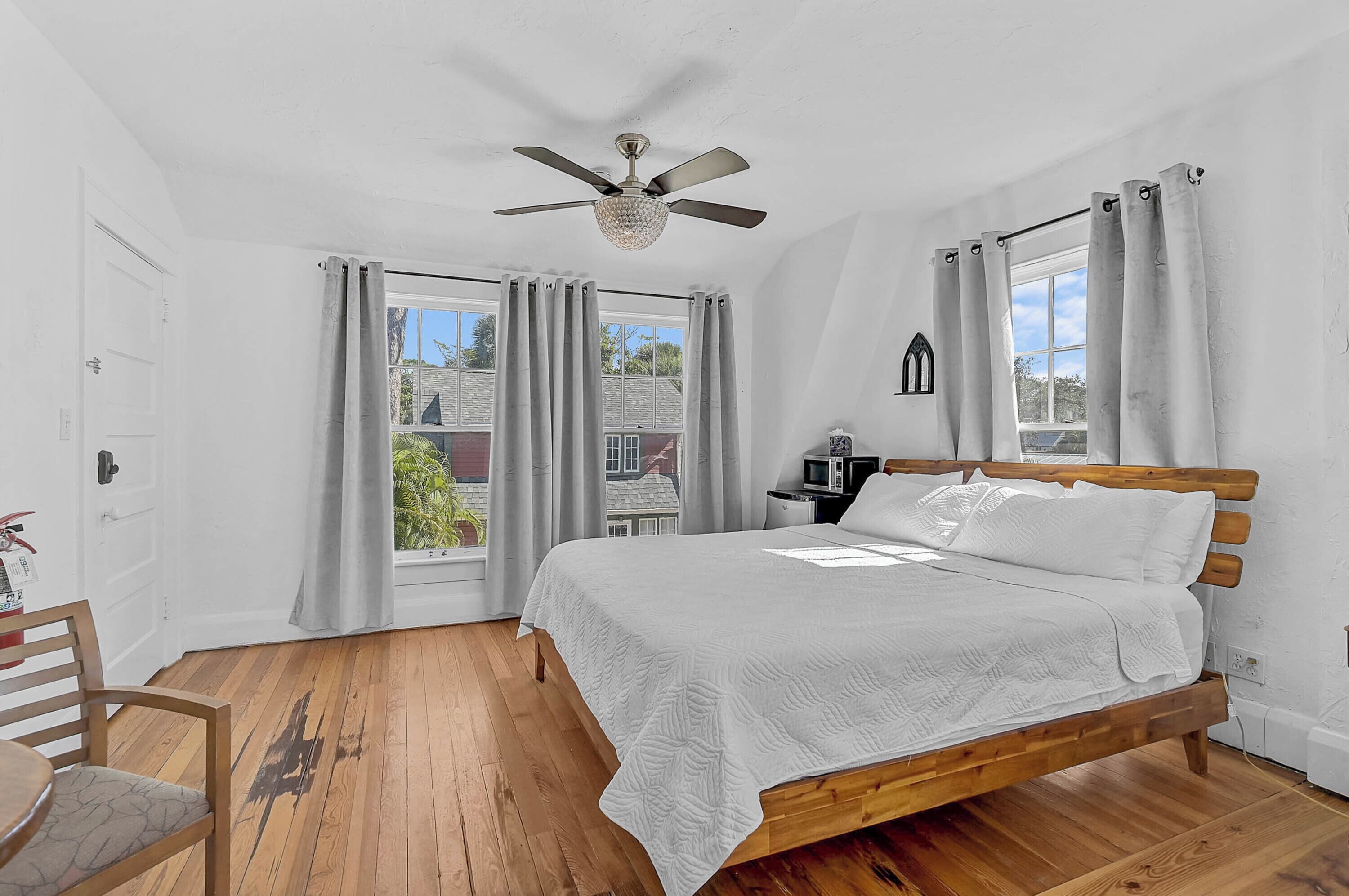 Un dormitorio con cama de madera, ventilador de techo, ventana con cortinas y vista a una casa.
