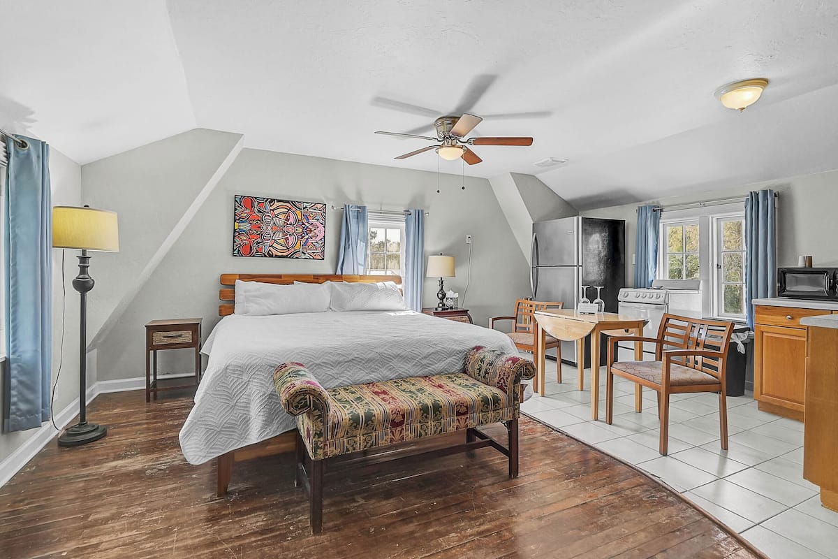 A bedroom with a bed, a bench, a desk, and a kitchenette.