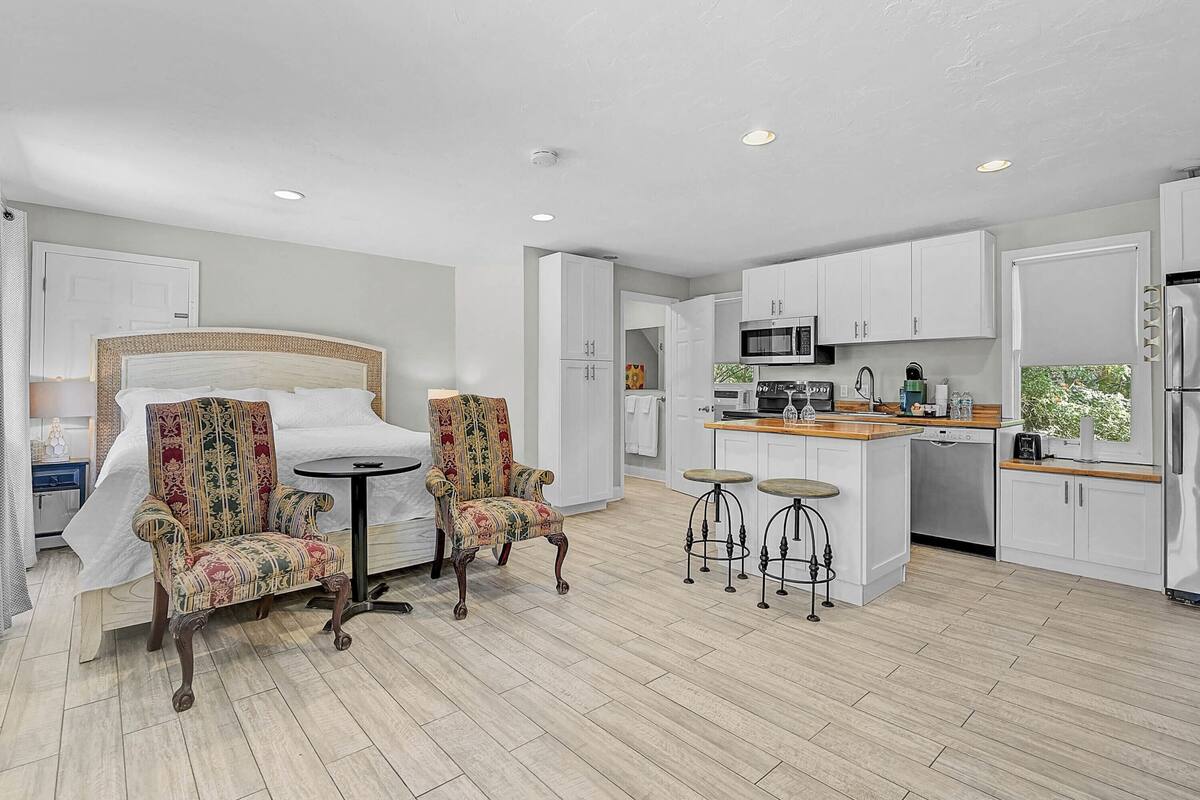 A modern hotel room with a large bed, two patterned armchairs, a small round table, and a kitchenette with a microwave, toaster, and refrigerator.