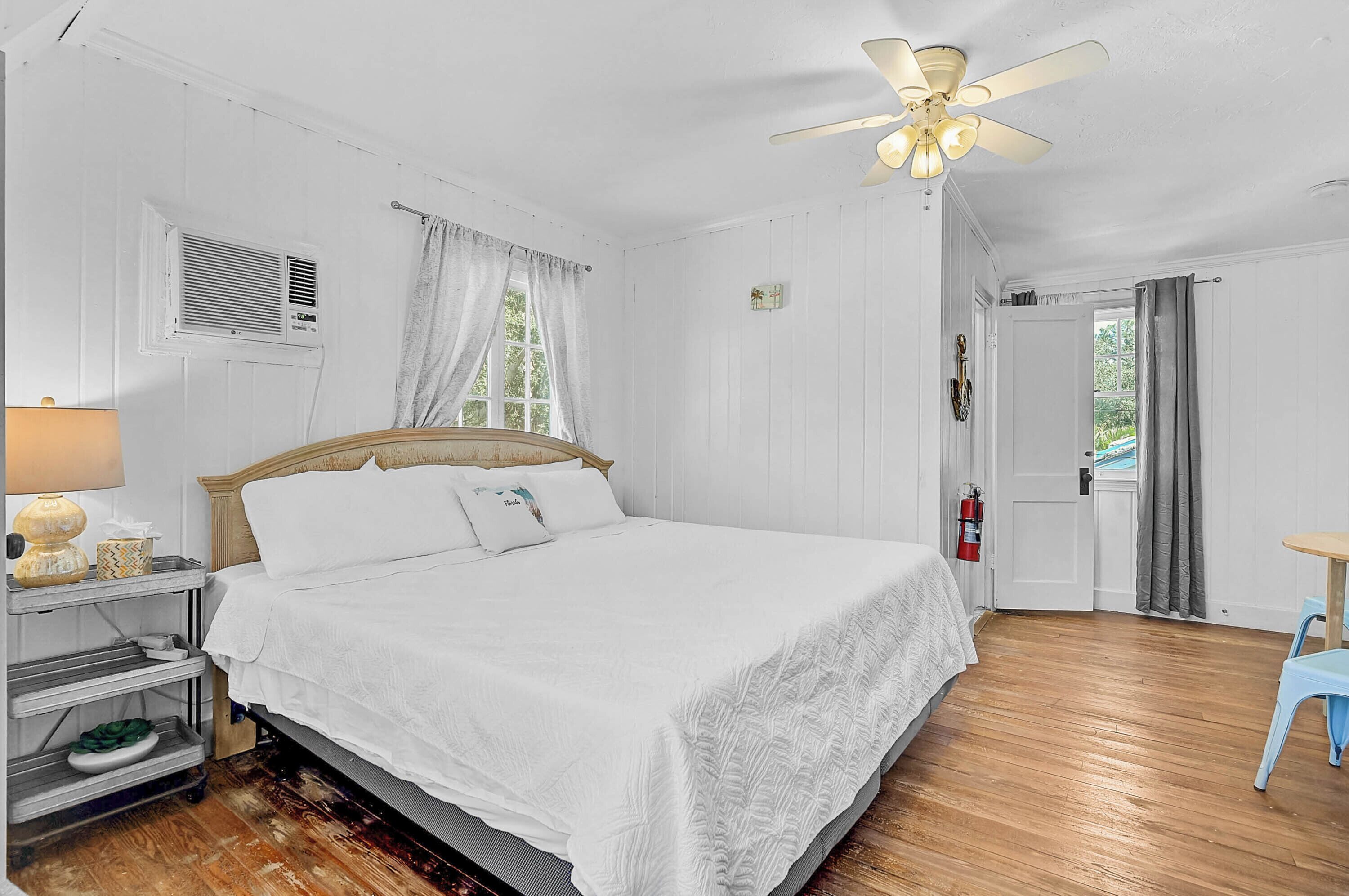 A bedroom with a wooden bed, white bedding, a ceiling fan, a window with curtains, a nightstand with a lamp, and a small table with a chair.