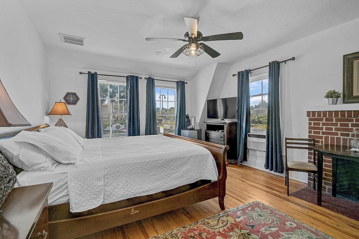 A bedroom with a bed, a desk, a chair, a TV, and a fireplace.