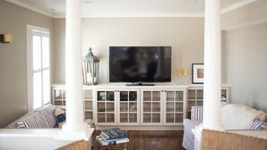 Un salon avec un grand téléviseur à écran plat posé sur un meuble blanc, des canapés à rayures et une petite table basse sur laquelle sont posés des livres.