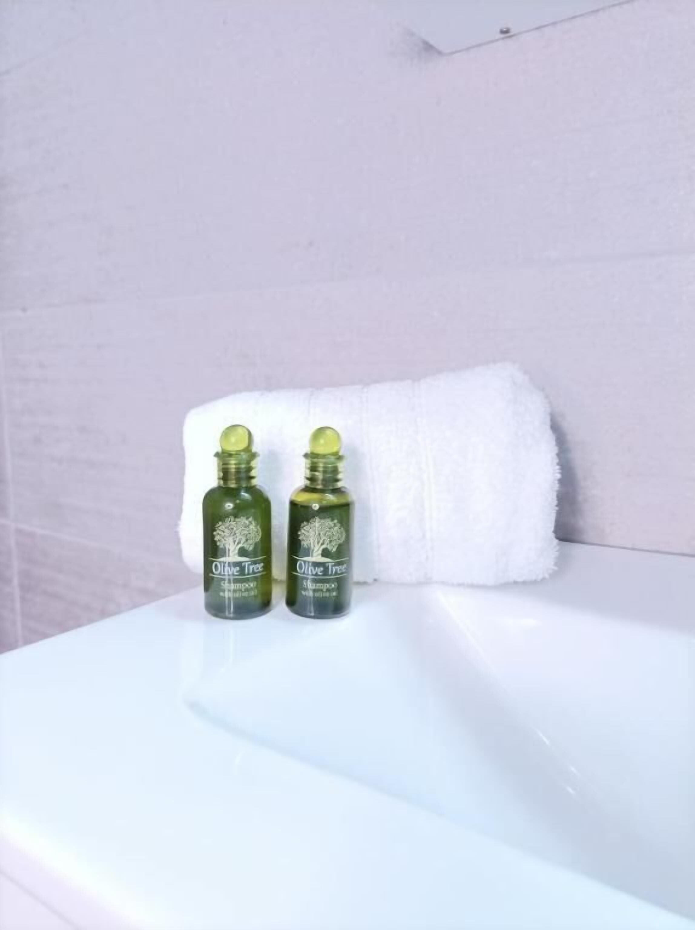 Two bottles of Olive Tree shampoo on a white bathroom counter with a white towel hanging on the wall.