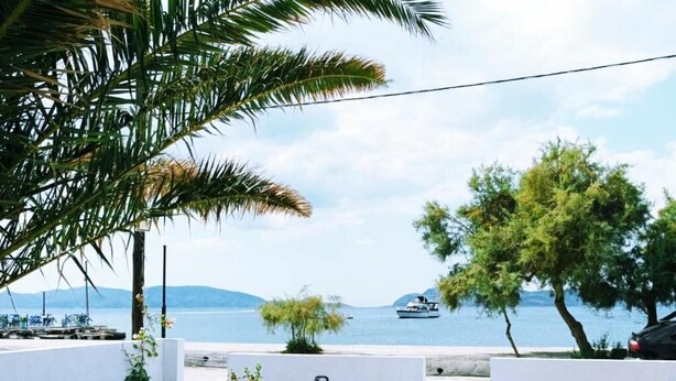 A coastal view with a white building, palm trees, and a boat in the distance. - V&E's Apartments (Ermionida)