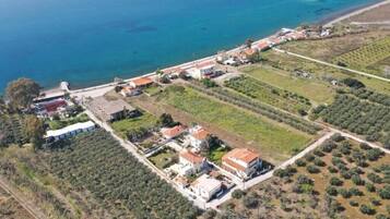 Vue aérienne d’une propriété côtière comprenant plusieurs bâtiments, notamment une grande villa avec un toit rouge, des constructions plus petites adjacentes et une plage en arrière-plan.