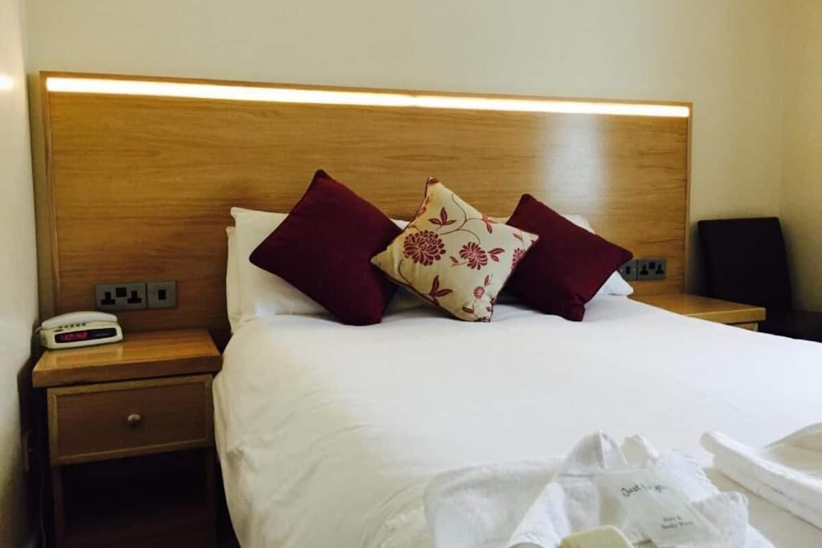 A hotel room with a wooden headboard, white bedding, two red pillows, a floral-patterned pillow, a bedside table with a clock, and a small drawer.