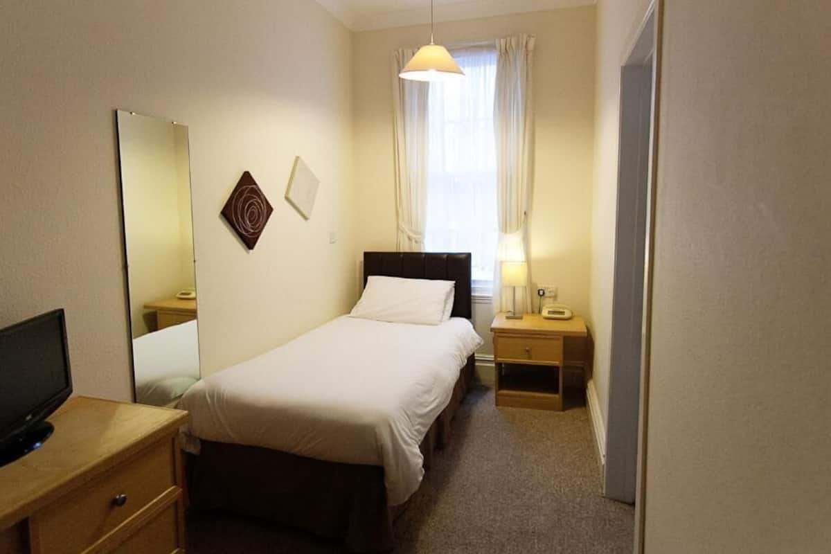 A hotel room with a single bed, a bedside table with a lamp, a TV on a wooden stand, a mirror, and a window with curtains.