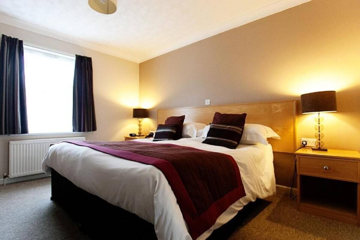 A hotel room with a large bed, two bedside tables with lamps, a window with curtains, and a radiator.