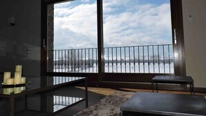 Ein Zimmer mit einer großen Schiebeglastür, die auf einen Balkon mit Blick auf ein Gewässer und einen bewölkten Himmel führt.