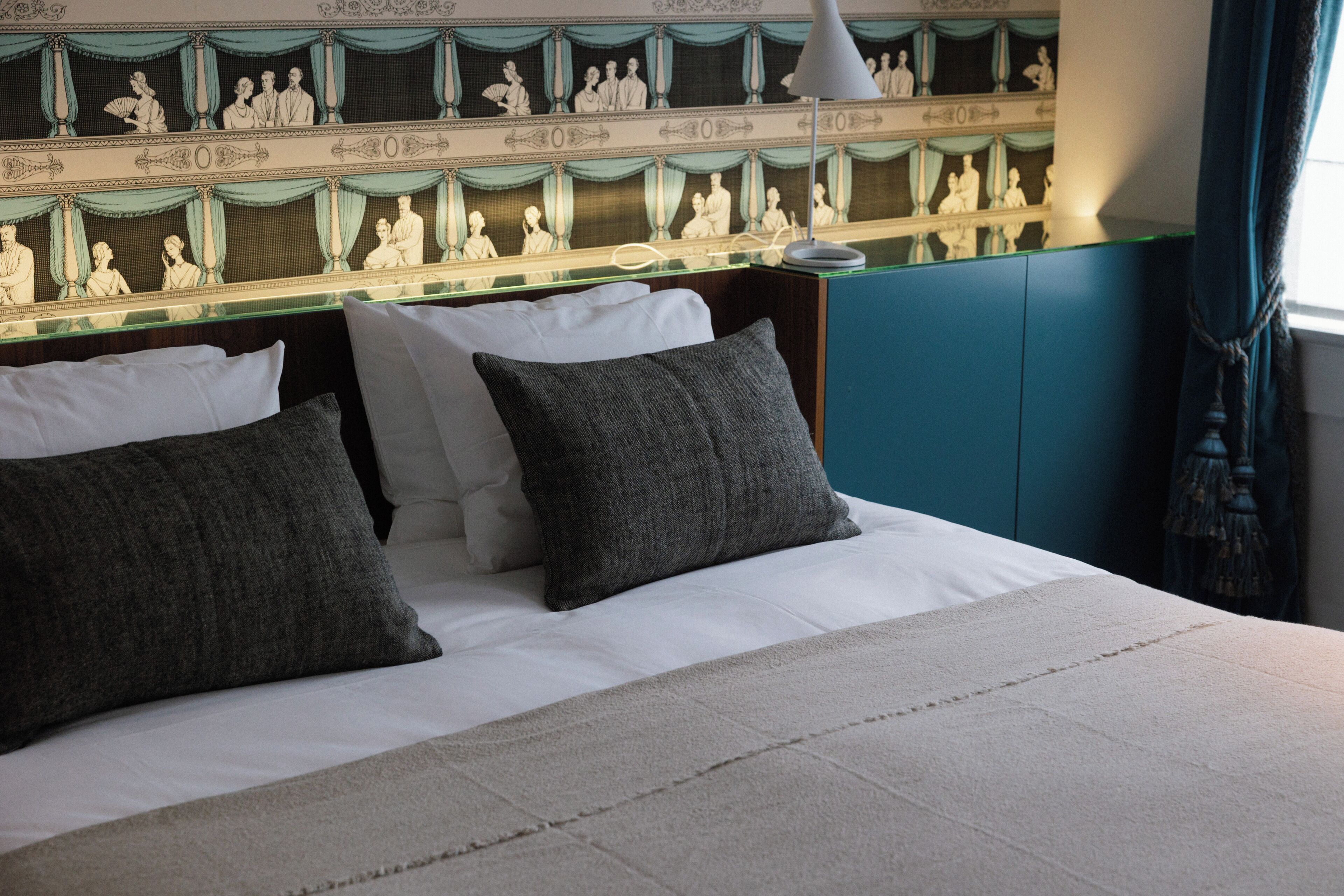Una cama bien hecha con sábanas blancas y dos almohadas oscuras, una lámpara de noche y un espejo decorativo en la pared que refleja una escena clásica.