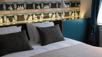 Una cama bien hecha con sábanas blancas y dos almohadas oscuras, una lámpara de noche y un espejo decorativo en la pared que refleja una escena clásica.
