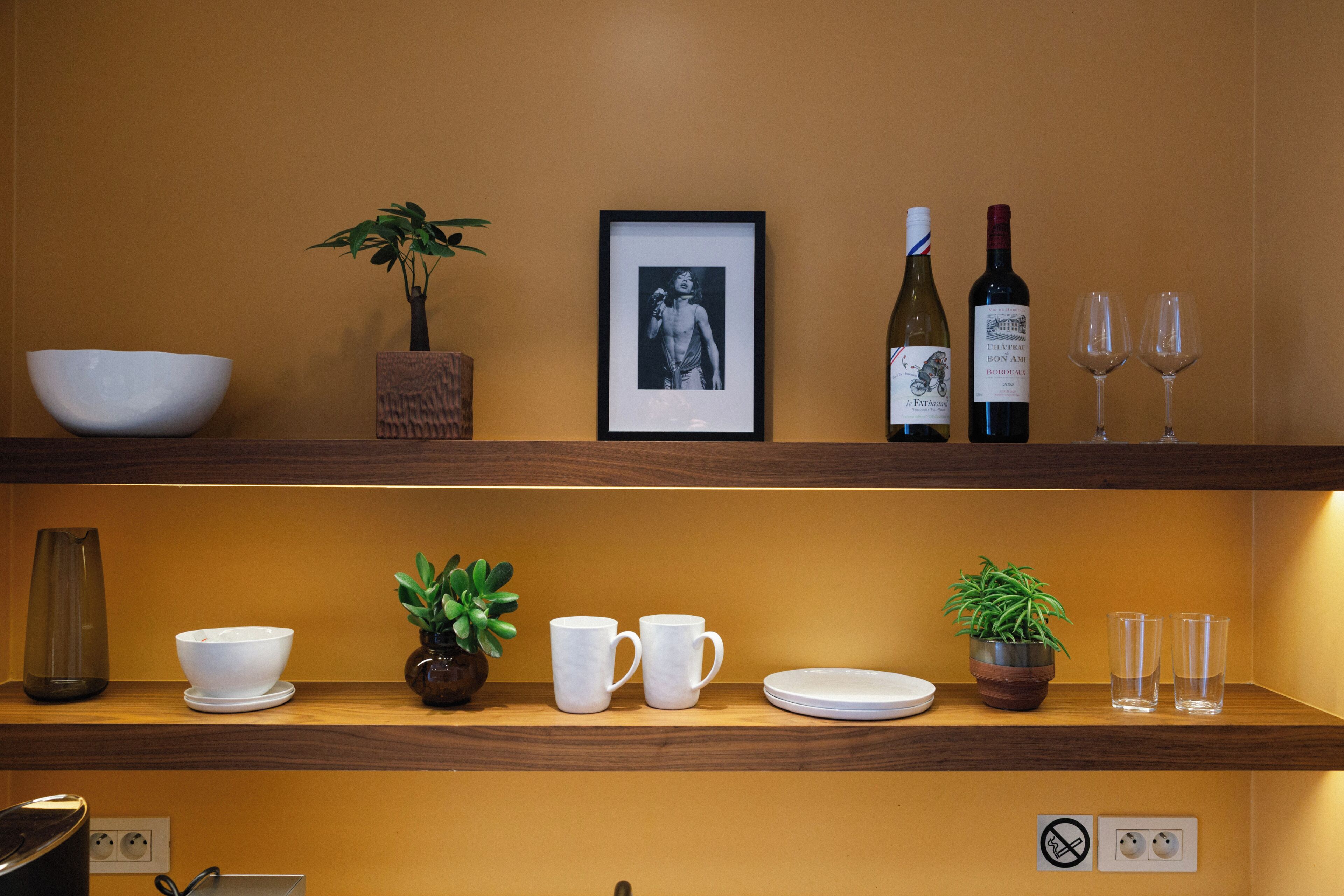 A shelf with a white bowl, two white mugs, a framed picture, a bottle of wine, and two wine glasses.