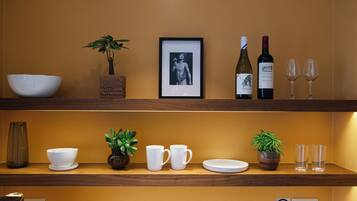 A shelf with a white bowl, two white mugs, a framed picture, a bottle of wine, and two wine glasses.