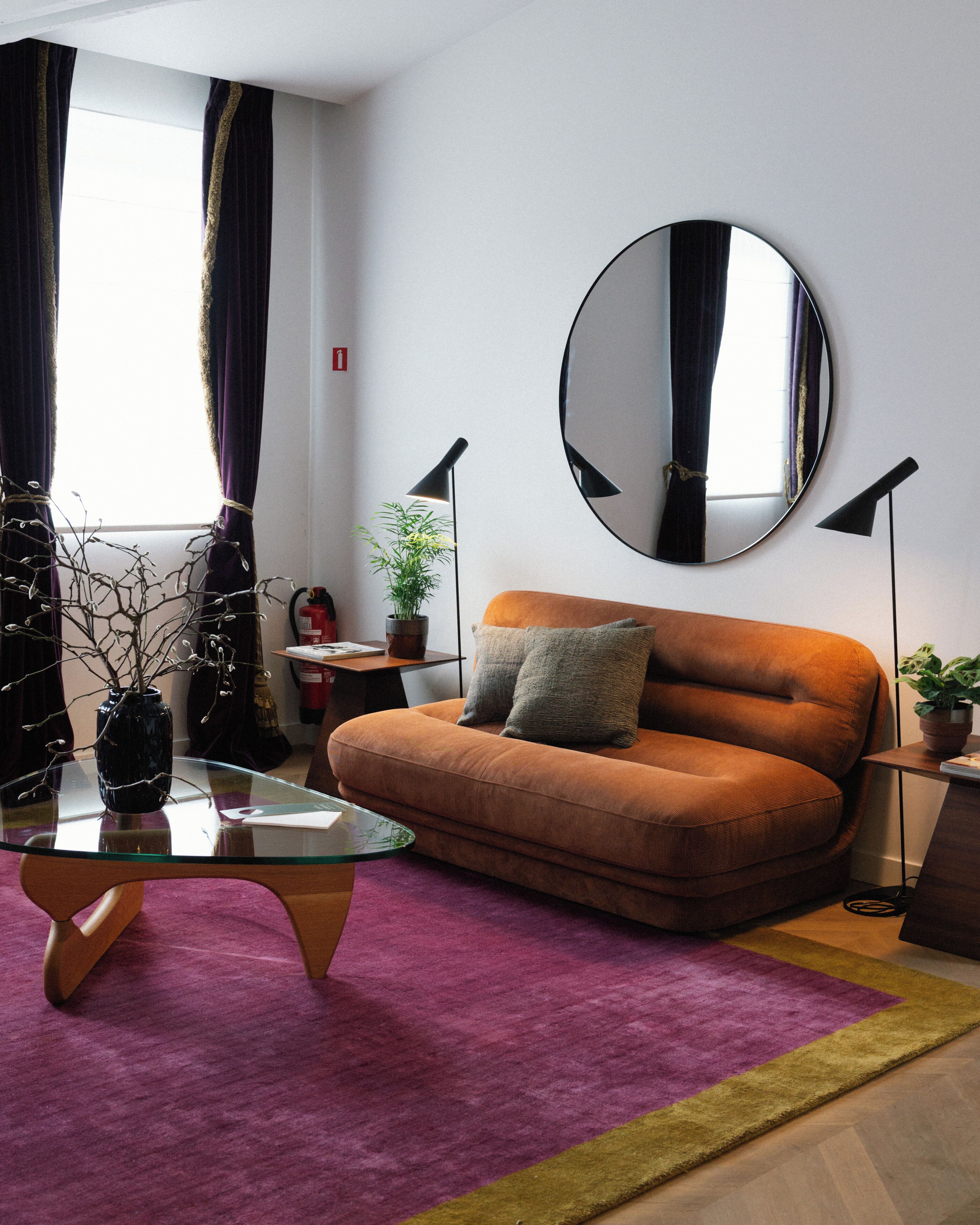 Ein modernes Wohnzimmer mit einem Glastisch, einer Couch mit Kissen, einem runden Spiegel an der Wand und einer Topfpflanze.