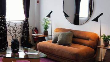 Ein modernes Wohnzimmer mit einem Glastisch, einer Couch mit Kissen, einem runden Spiegel an der Wand und einer Topfpflanze.