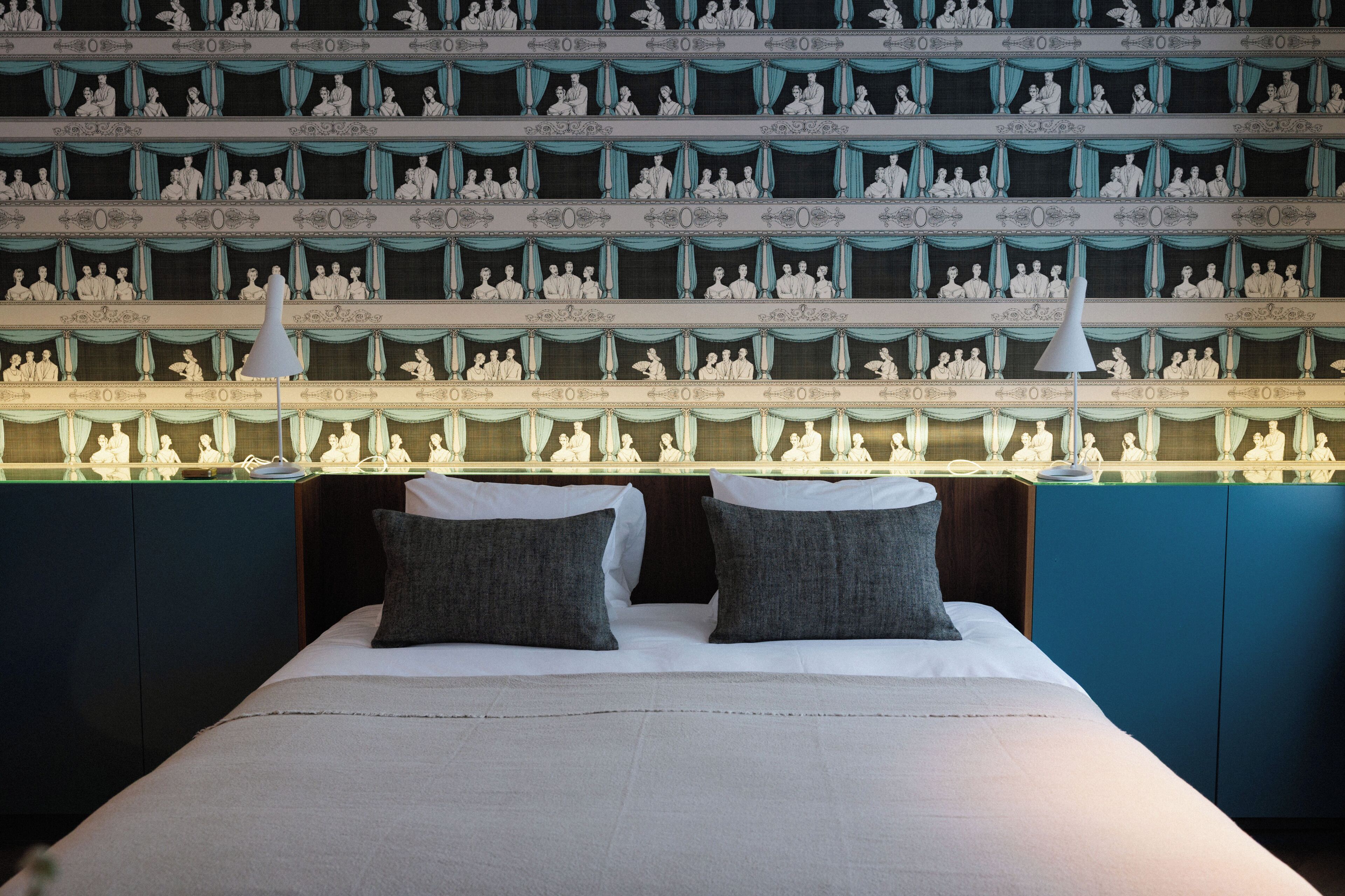 Habitación de hotel con una cama grande, dos almohadas, una mesita de noche y una pared con múltiples retratos enmarcados.