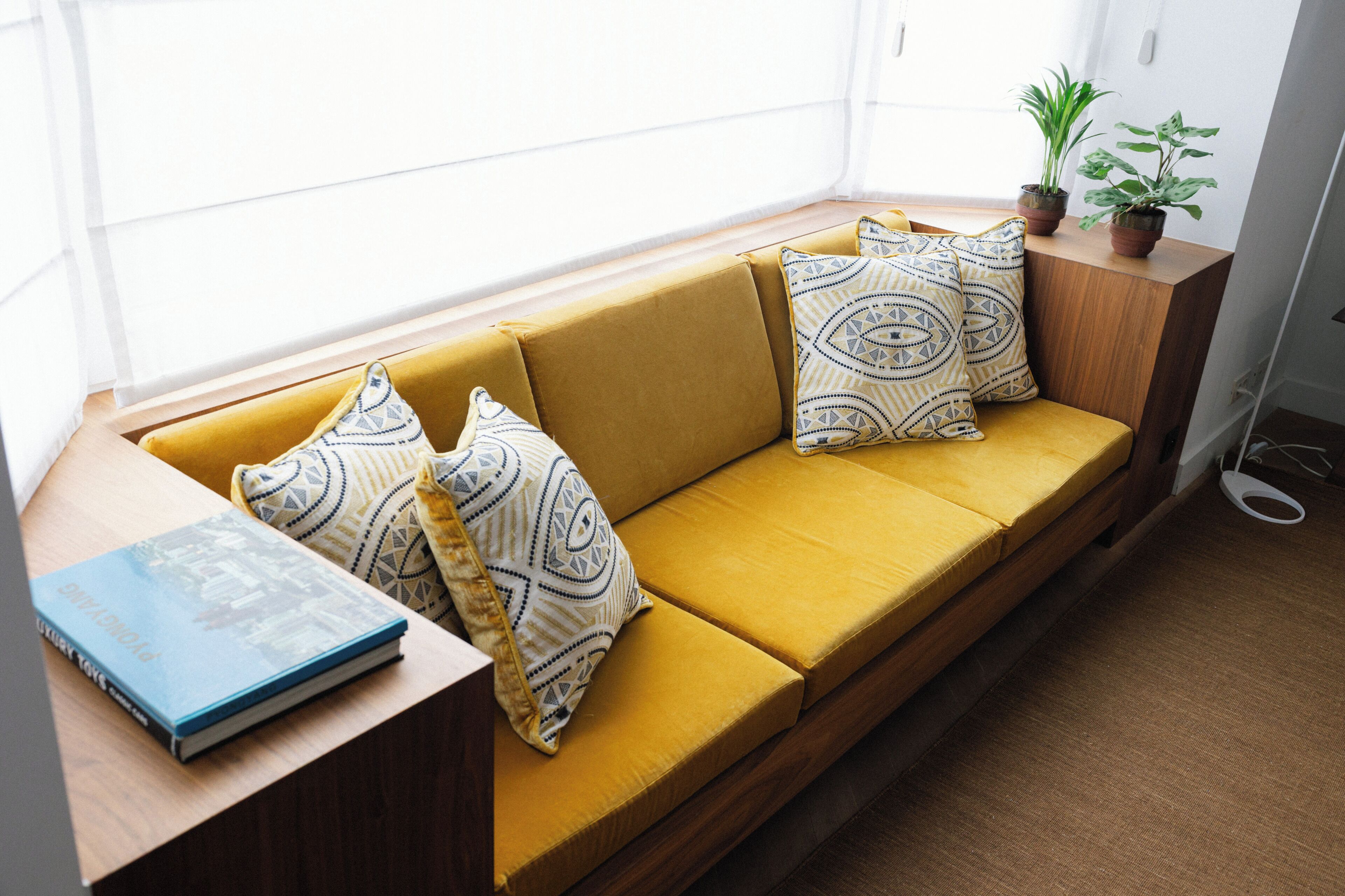 A yellow sofa with patterned cushions, a book on a wooden side table, and a potted plant on a windowsill.
