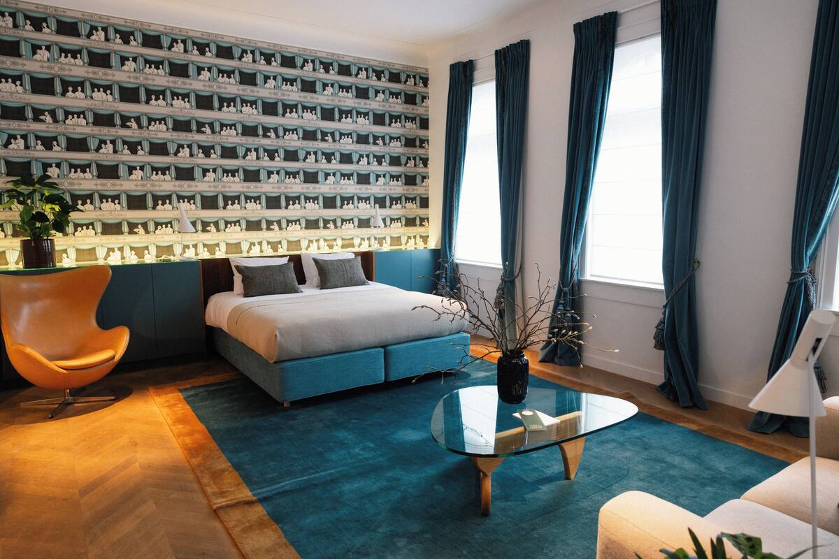 Une chambre moderne avec un lit, un canapé bleu, une table basse en verre et un mur recouvert d’étagères décoratives.