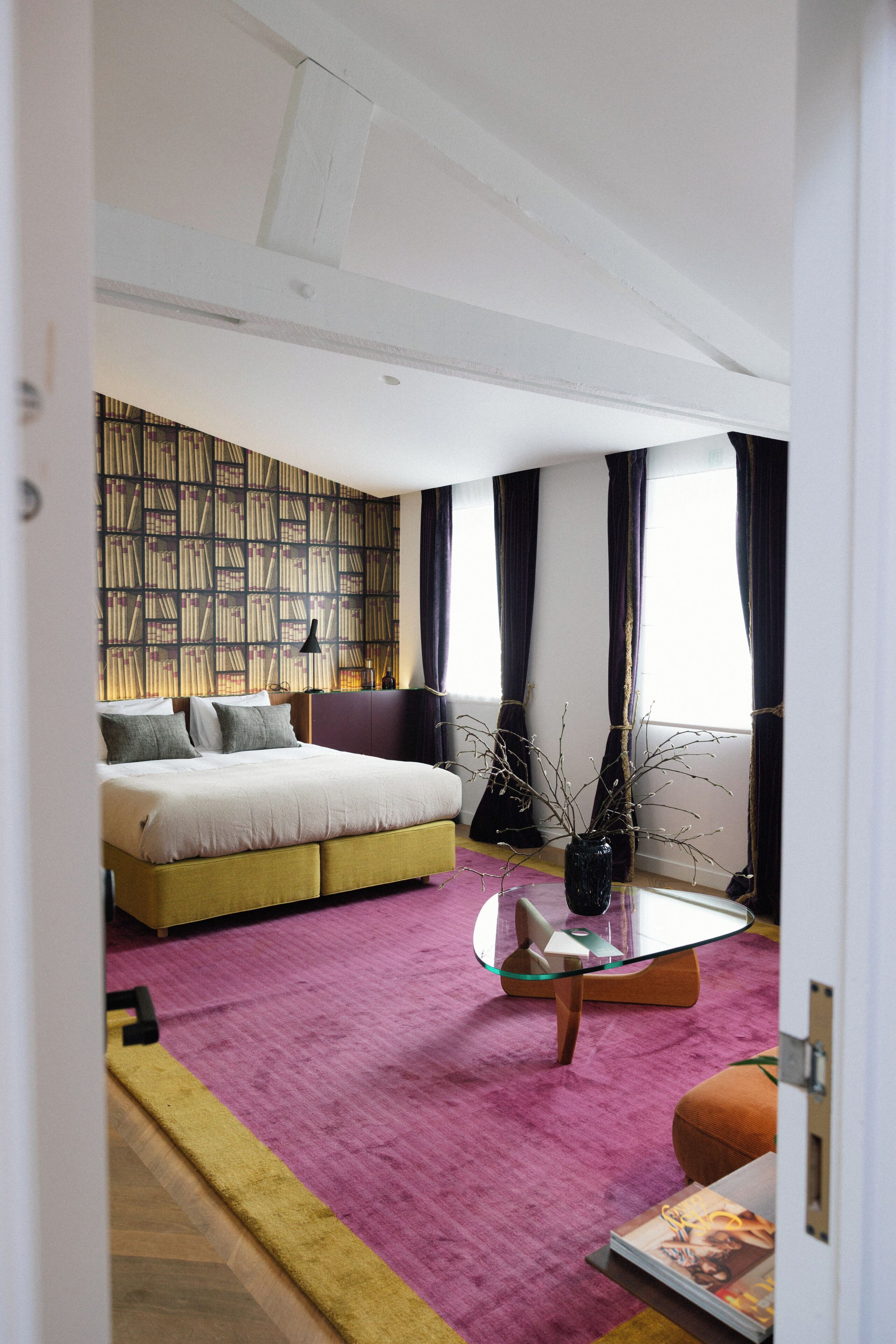 Un dormitorio moderno con una cama, una mesa de centro de vidrio, una alfombra morada y una estantería.