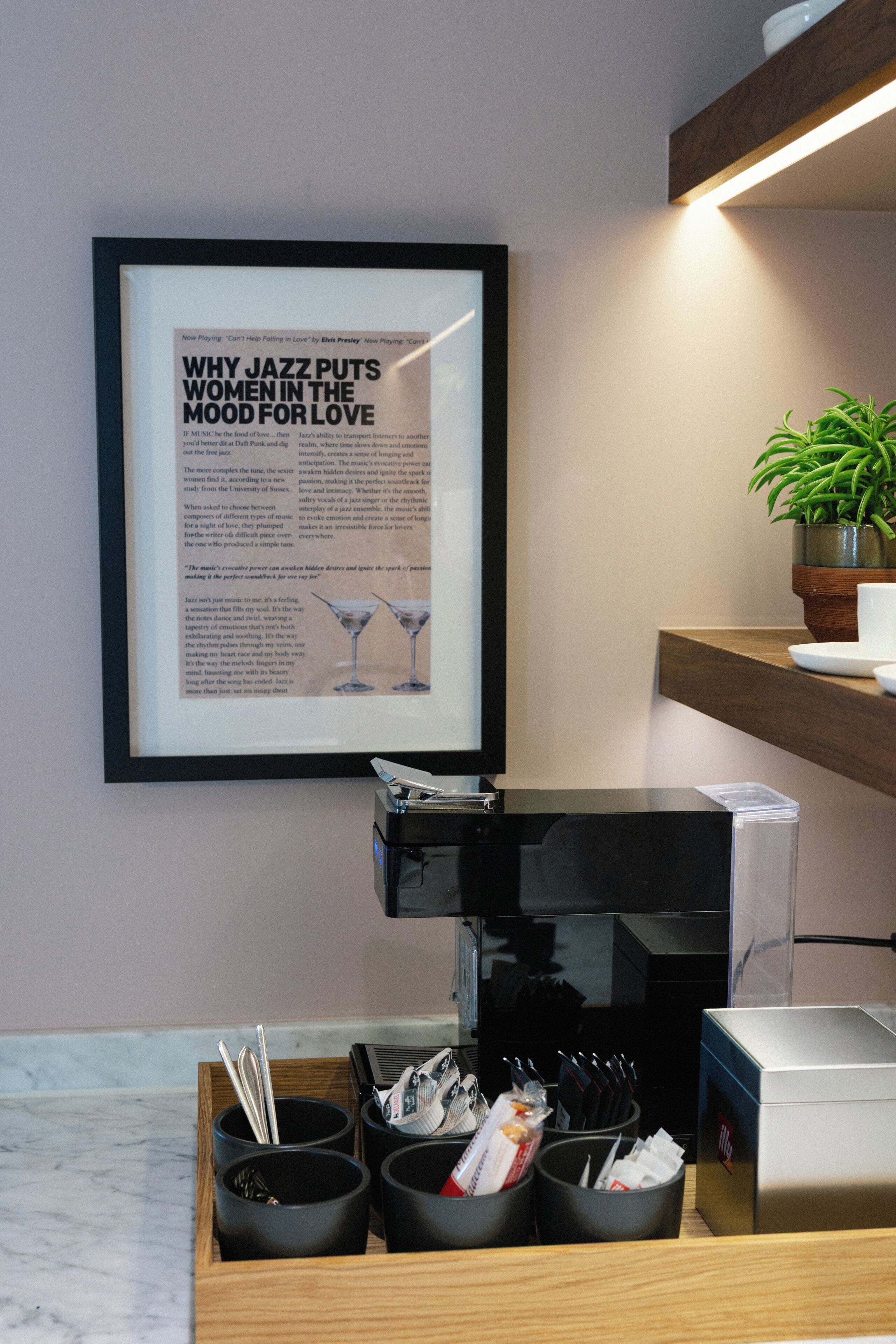 A framed article on a wall, a coffee machine, a tray with cups and utensils, and a potted plant.