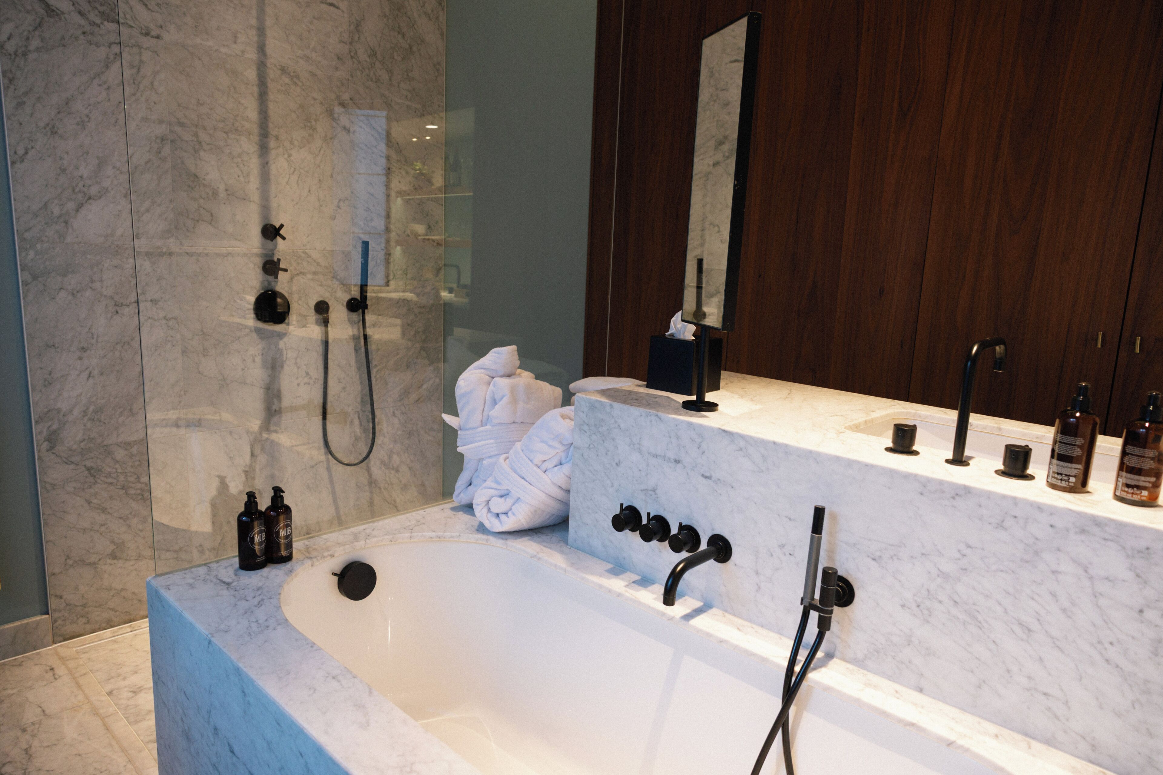 A modern bathroom with a marble bathtub, a large mirror, and a wooden vanity.