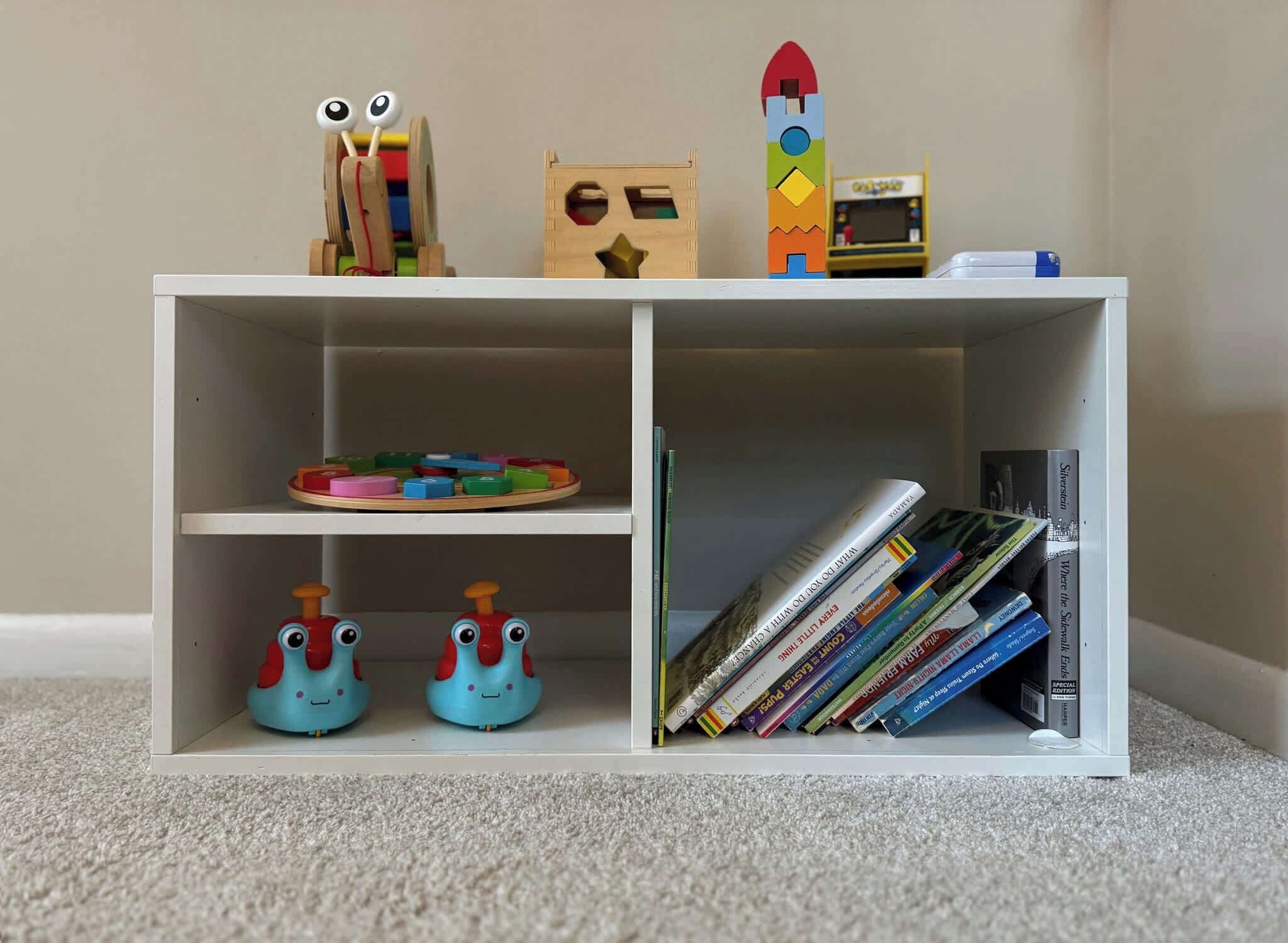 Una estantería blanca con dos niveles; la repisa superior alberga juguetes de madera y un pequeño cohete de juguete, la repisa central tiene bloques de construcción de colores, y la repisa inferior contiene dos coches de juguete azules y una pila de libros.