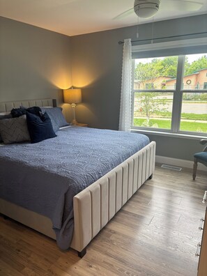 A bedroom with a large bed, a bedside lamp, a window with curtains, and a view of the outside. - Walking distance to local eateries, watering holes, beaches, grocery & shopping! (Elk Rapids)