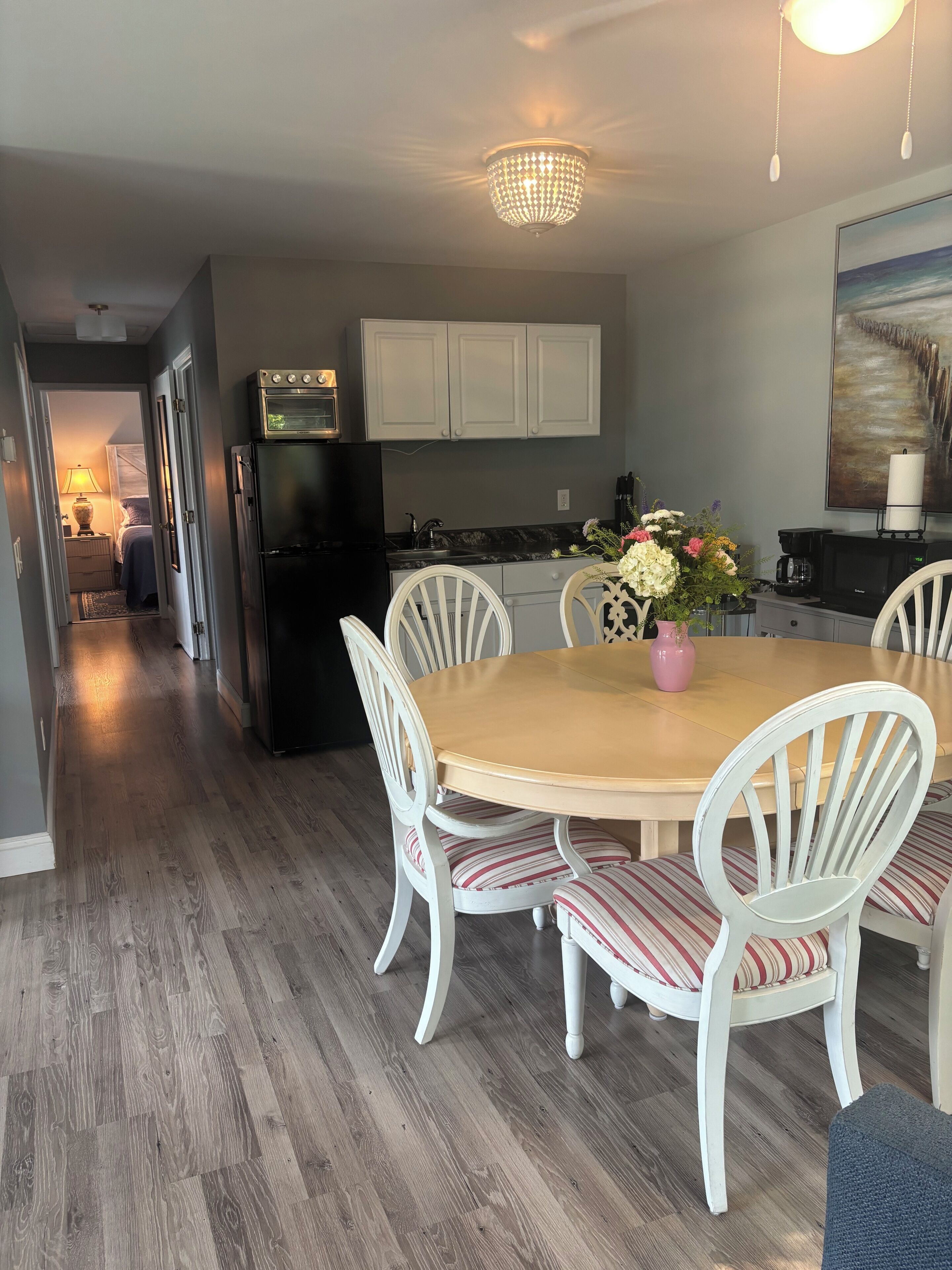 A dining area with a wooden table, white chairs, a chandelier, and a kitchen in the background.
