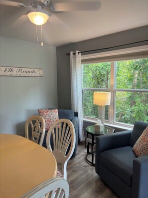 A room with a dining table, two chairs, a sofa, a lamp, and a window with a view of greenery. - Walking distance to local eateries, watering holes, beaches, grocery & shopping! (Elk Rapids)