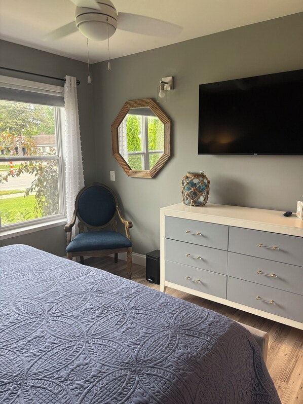 Un dormitorio con cama, una silla, cómoda, televisor y ventana con vista a follaje.