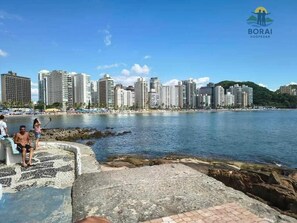 A coastal cityscape with high-rise buildings, a rocky shoreline, and a clear blue sky. - Apartment with 1 bedroom and balcony, sea view. (Guarujá)