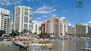 Un paisaje urbano costero con rascacielos, una playa con gente y barcos, y un cielo despejado.