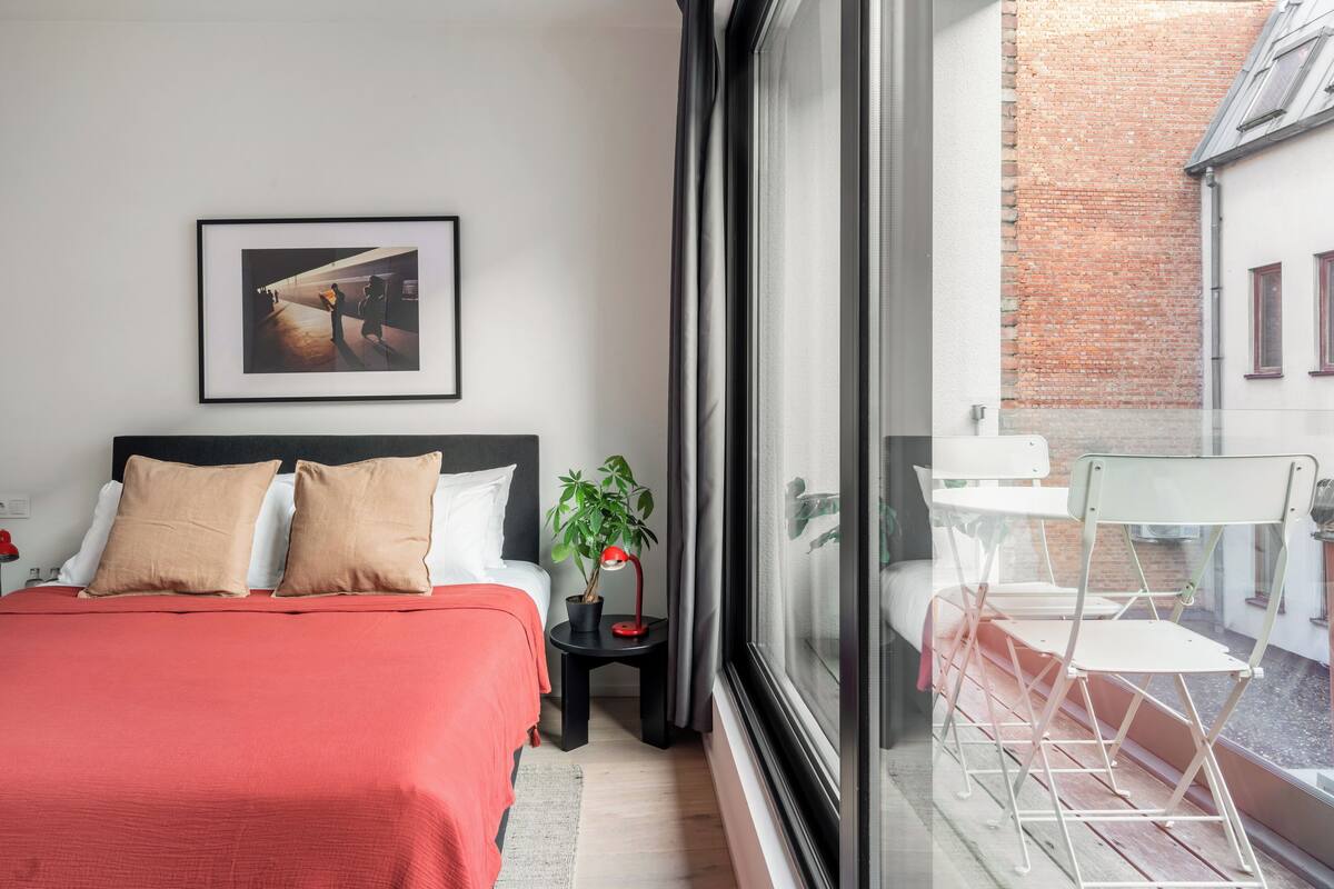 Een hotelkamer met een bed, een lijstbeeld aan de muur, een tafeltje met een plant en een balkon met stoelen.