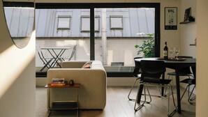 Een moderne woonkamer met een bank, een bijzettafeltje met boeken, een eethoek met een tafel en stoelen, en een groot raam met uitzicht op gebouwen.