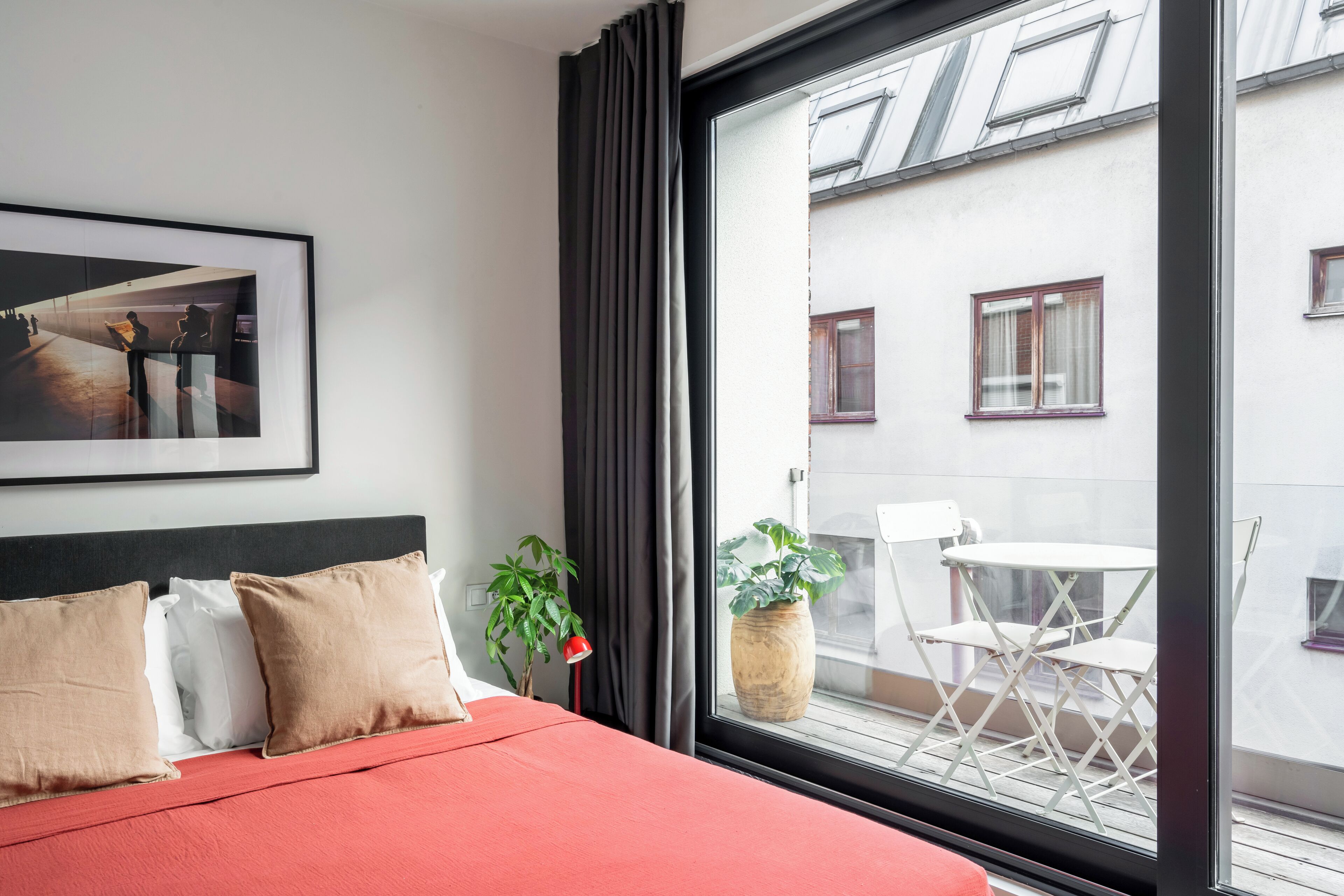 Una habitación de hotel con un ventanal grande, una cama con ropa de cama roja, un cuadro en la pared y una planta en maceta.