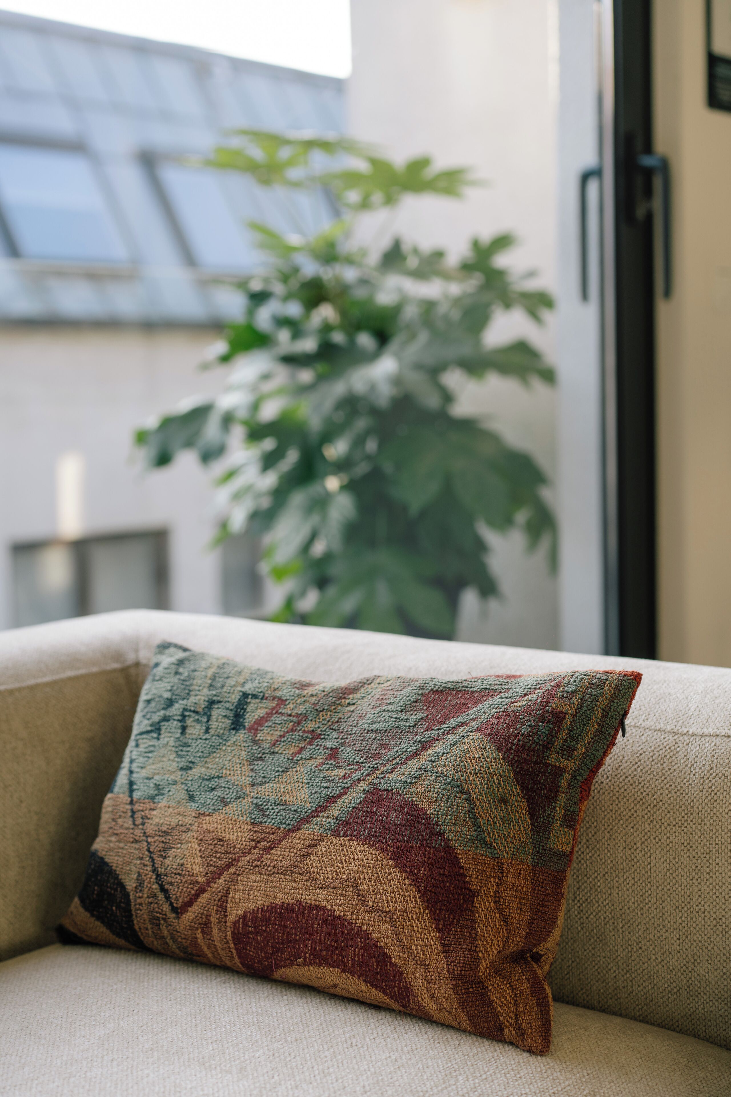 A beige sofa with a patterned throw pillow, a potted plant, and a window with a view of a building.