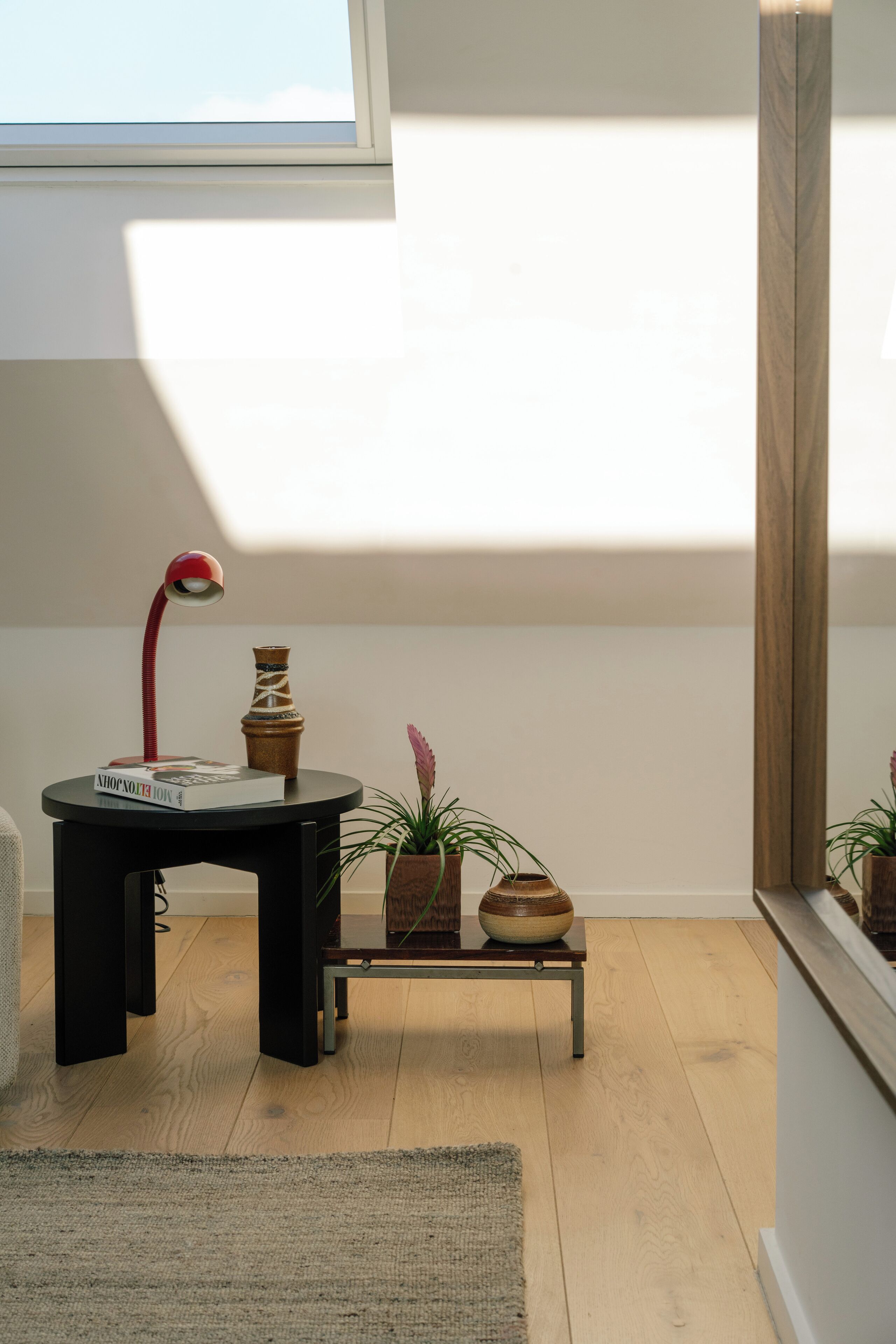 Ein Zimmer mit Holzboden, einem schwarzen Tisch mit roter Lampe, einer Vase und einem kleinen Regal mit Pflanzen.