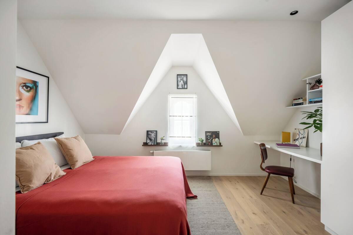 Een slaapkamer met schuine wand, een bed met rode beddengoed, een bureau met stoel en een raam met uitzicht.