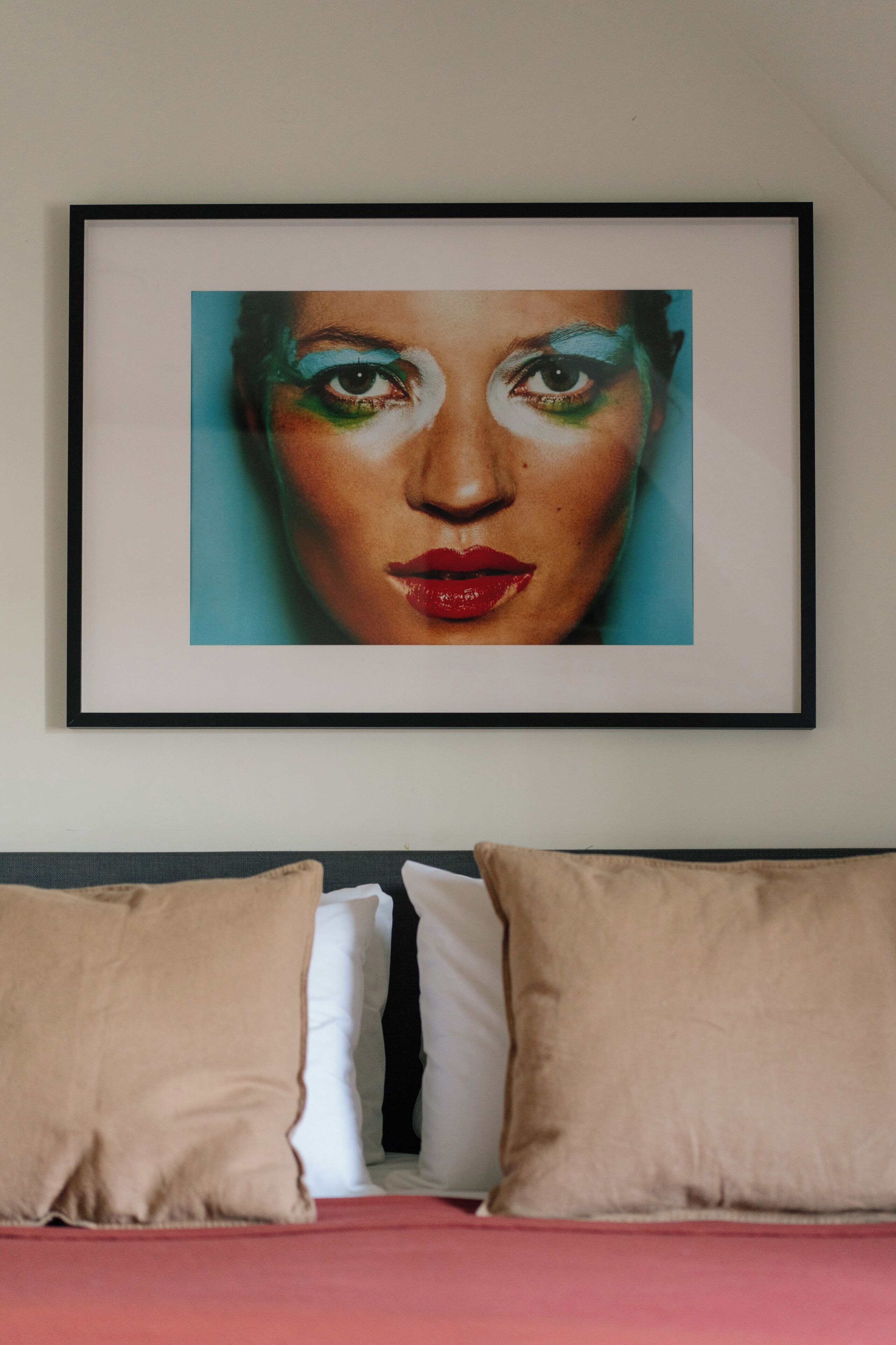 A framed artwork on a wall above a bed with beige pillows and a red bedspread.