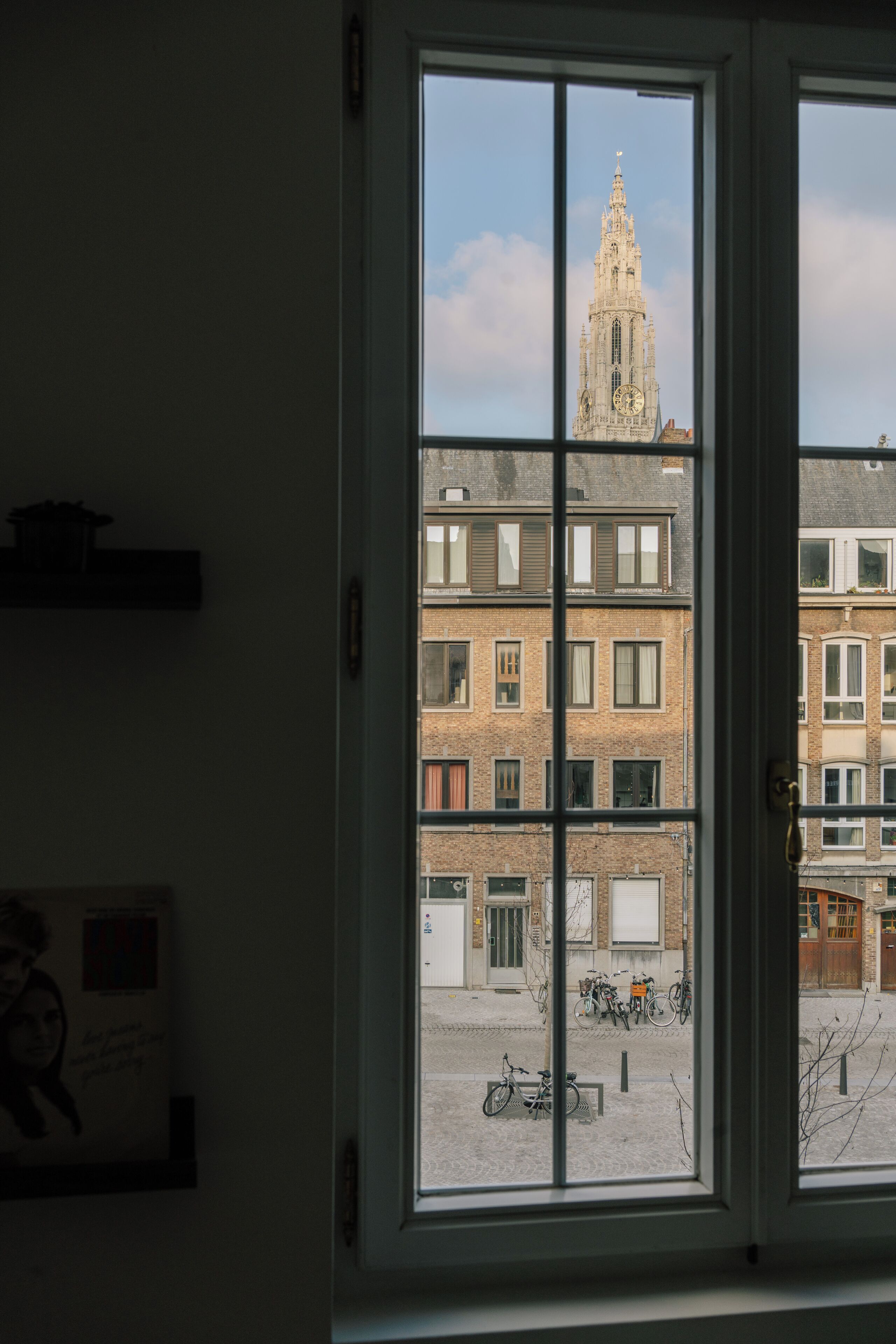 Una vista attraverso una finestra: un edificio con una torre dell'orologio, biciclette e una strada acciottolata.