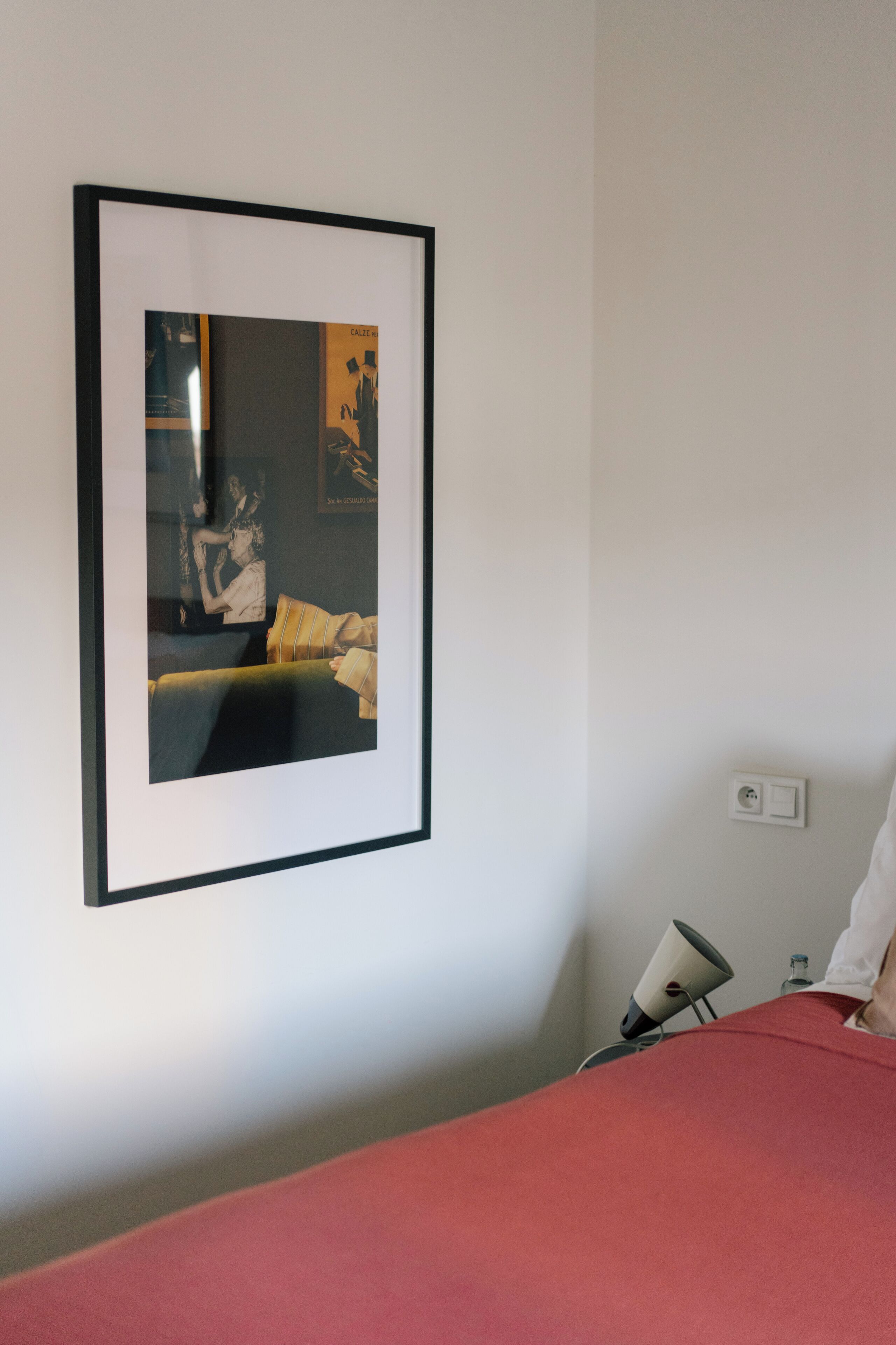Een lijst met een foto aan een muur, een bed met een rode spreidek en een lamp op een nachtkastje.