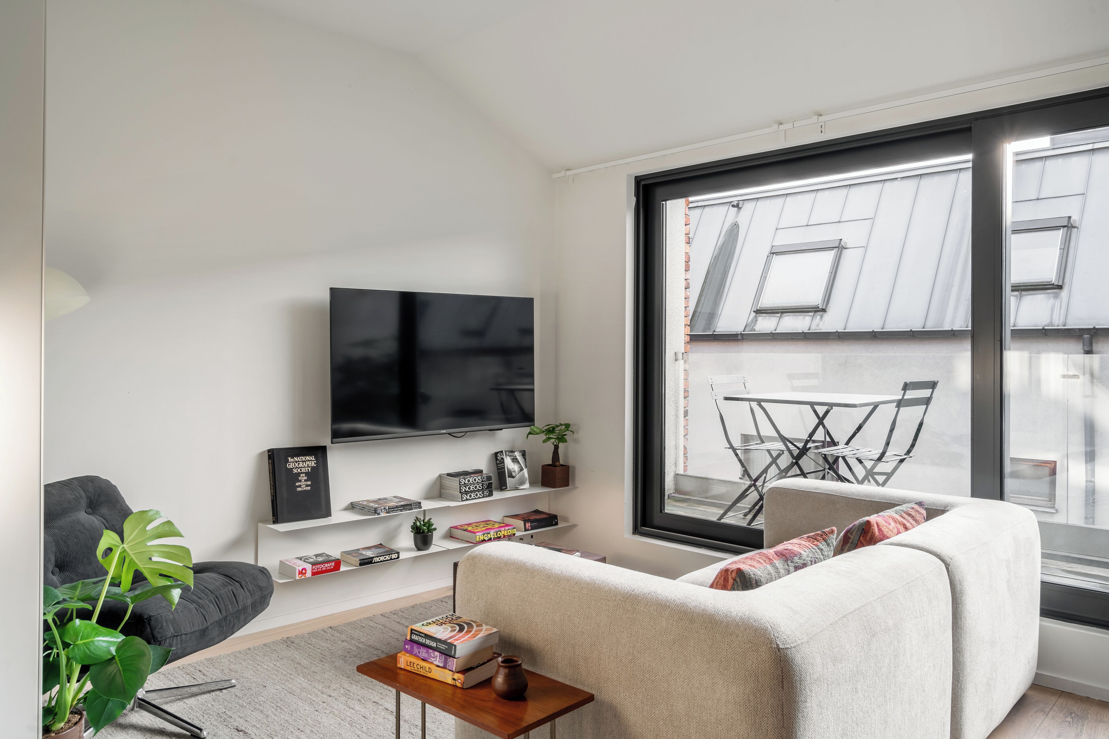 A modern living room with a flat-screen TV, a grey sofa, a white sofa, and a large window with a view of a building.