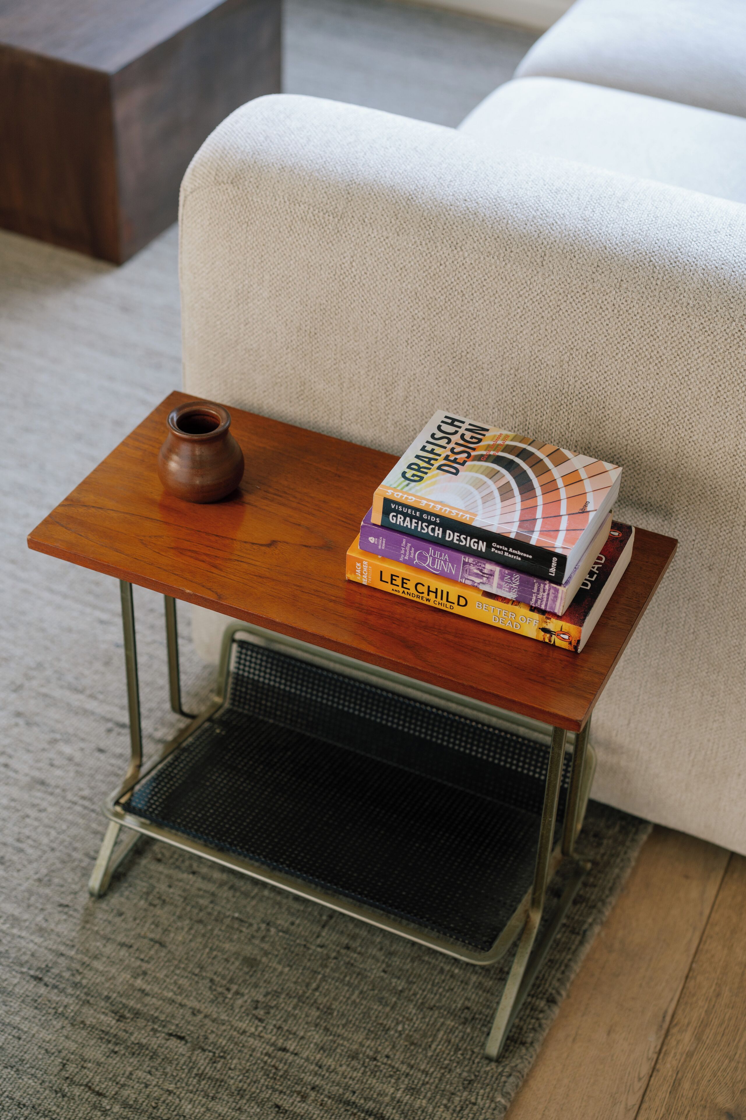 A small wooden side table with a metal base, a brown ceramic pot, and several books including titles like 'GRAFISCH DESIGN'.