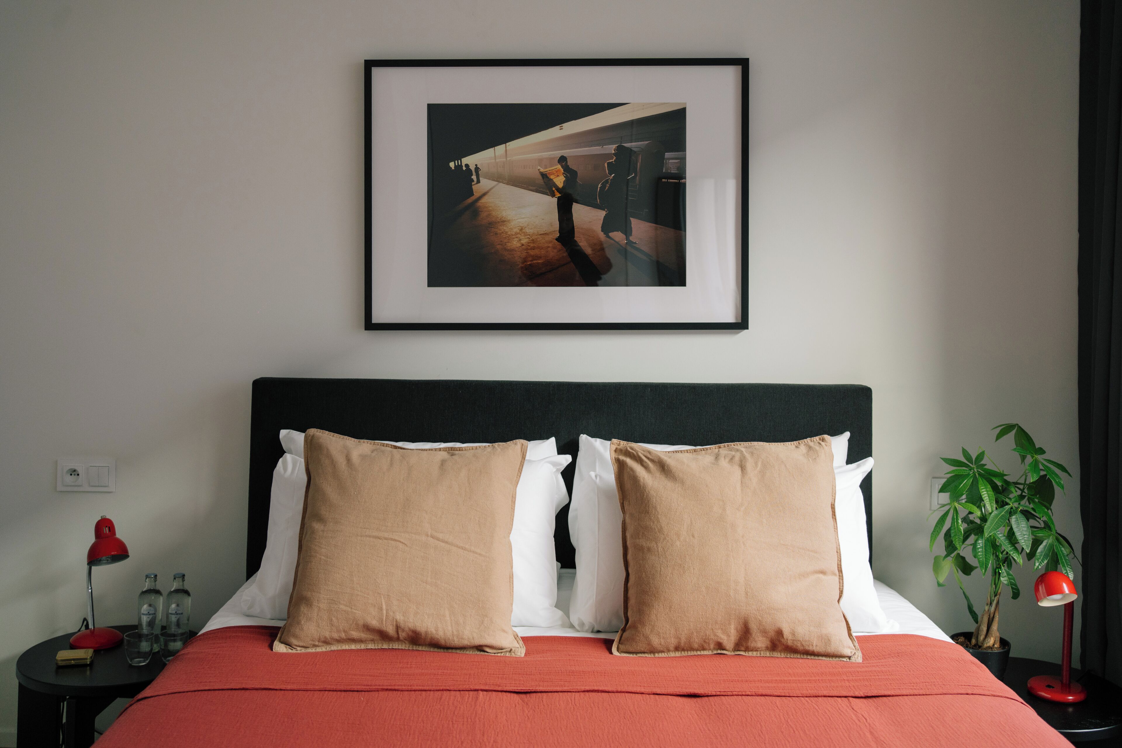 A neatly made bed with two pillows, a framed picture above it, a nightstand with a lamp, and a potted plant.