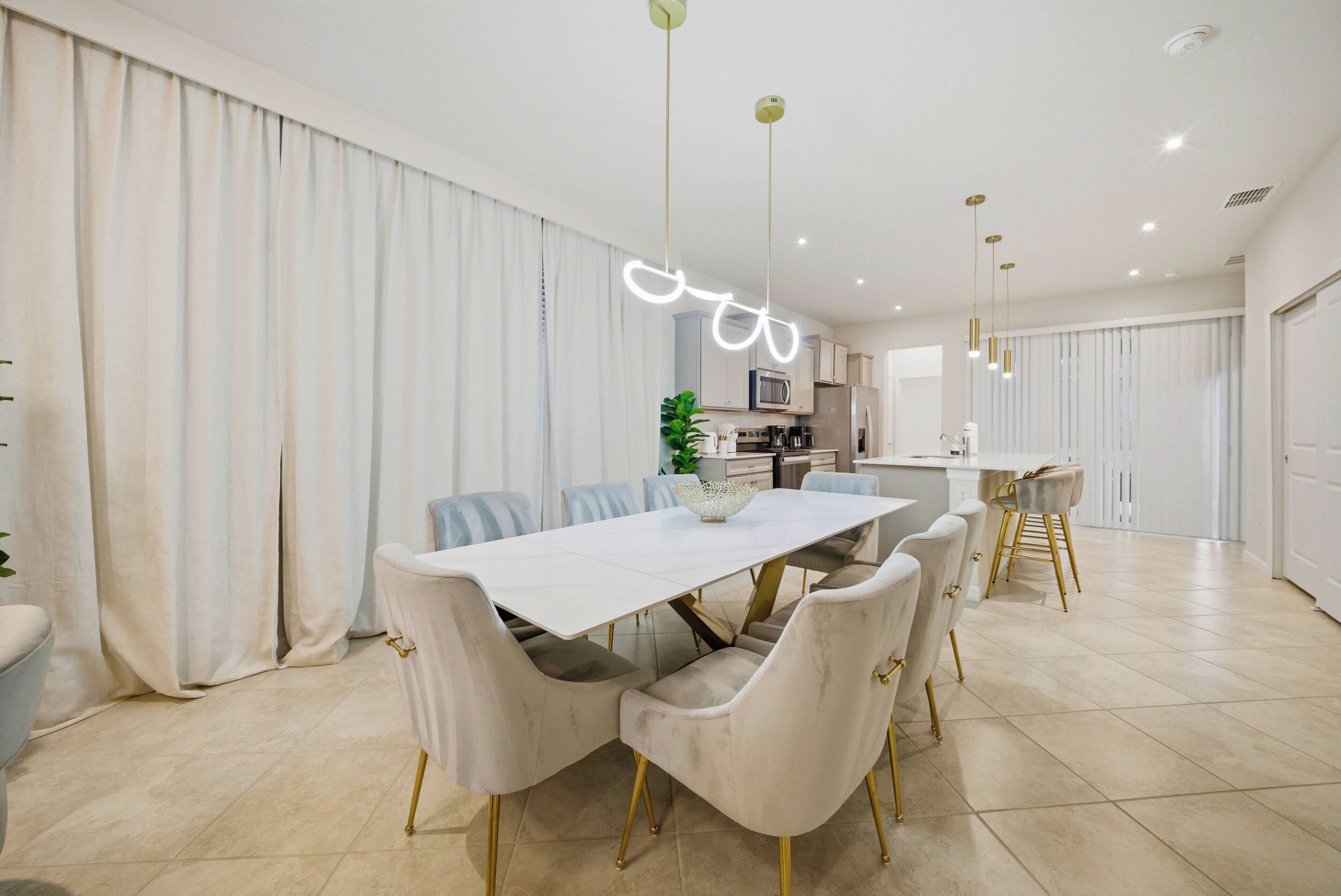 A modern dining area with a white table and matching chairs, a kitchen in the background, and a bright pendant light.
